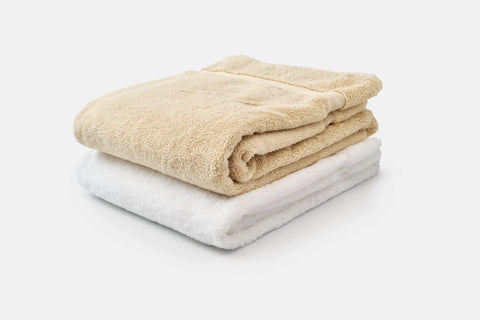 A beige Bath Towel Made of Luxury USA Cotton by American Blossom Linens rests atop a white one, both crafted from premium cotton for softness, neatly stacked against a plain white background.
