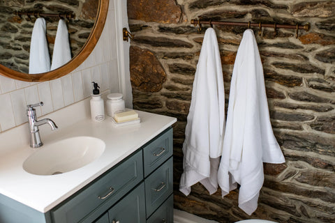 A bathroom with a stone wall, two American Blossom Linens Bath Towels Made of Luxury USA Cotton (700 GSM) in white on a copper bar, a round mirror, and a white sink on a blue vanity with soap, lotion dispenser, and cup on the counter.