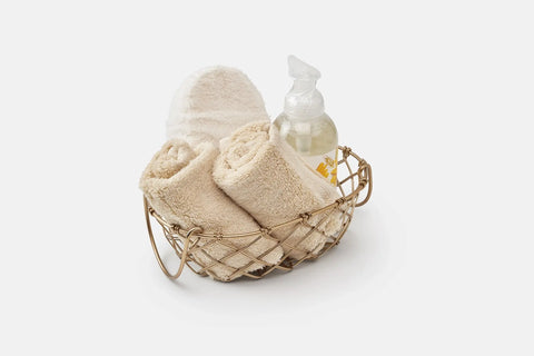 A gold wire basket holds two American Blossom Linens Bathroom Washcloths made of Luxury USA Cotton, white slippers, and a clear pump bottle of soap, perfect for an indulgent self-care ritual on a pristine white background.