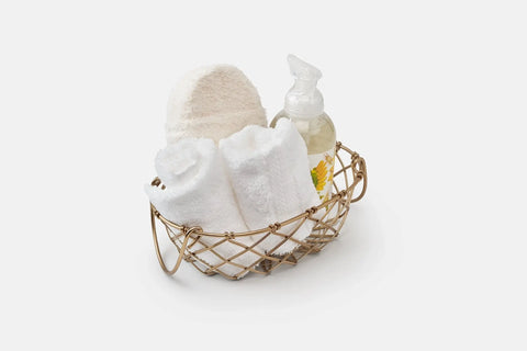 A gold wire basket holds two Bathroom Washcloths Made of Luxury USA Cotton from American Blossom Linens, a white textured wash mitt, and a floral-design clear soap bottle—an ideal self-care set.
