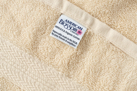 A close-up of a beige Bathroom Washcloth by American Blossom Linens, crafted from luxury USA cotton, featuring a label that reads: “100% U.S.A. Organic Cotton. Incredibly made in the U.S.A. by your friends & neighbors.”.