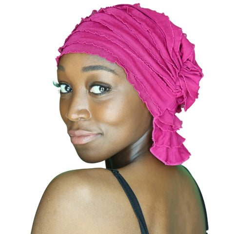 A woman with smooth dark skin wears the bright pink Chemo Beanies® Kathryn scarf and a black shoulder strap, looking over her shoulder at the camera with a soft smile against a white background.