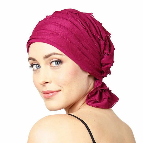 A woman with fair skin and blue eyes smiles gently over her shoulder, wearing the Chemo Beanies® Kathryn scarf—a bright magenta textured chemo beanie tied at the back. The background is plain white.