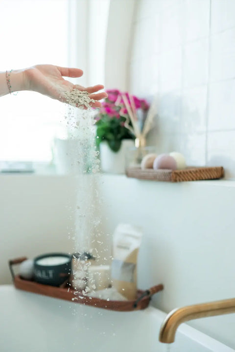 A hand sprinkles Shanti + Tula Magnesium Detox Bath Soak with Lavender & Bentonite Clay into a white tub, while pink flowers, bath bombs, and spa essentials on a shelf create a tranquil atmosphere for deep relaxation and sleep support.