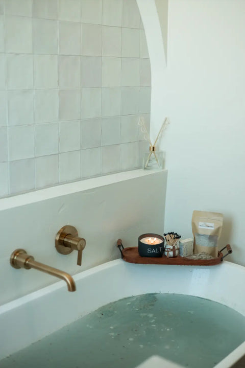 A modern tub with light blue water invites relaxation, featuring a wooden tray holding a lit candle, Shanti + Tula Magnesium Detox Bath Soak with Lavender & Bentonite Clay, bath products, and a diffuser against white tiles and a gold faucet.