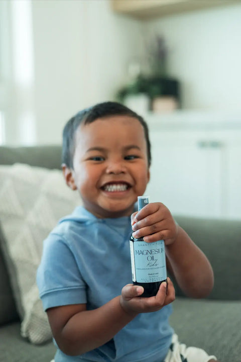 A smiling child in a blue shirt sits on a gray couch, holding Shanti + Tula Magnesium Oil Spray for Sleep, Stress & Muscle Recovery. The bright room with white cabinets and a plant highlights this pure, USA-made topical body oil.