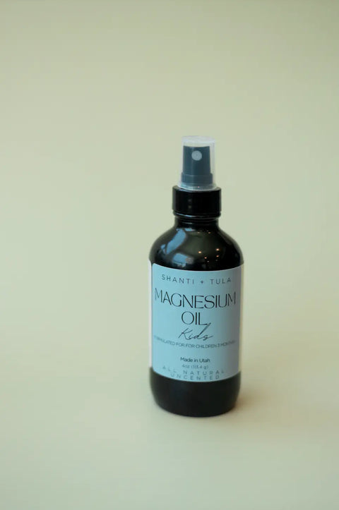 A dark spray bottle of Shanti + Tula Magnesium Oil Spray for Sleep, Stress & Muscle Recovery sits on a light surface; the pure, USA-made topical oil is formulated to support restful sleep and ease muscle soreness.
