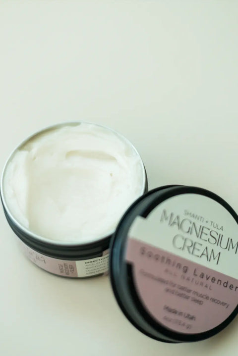 An open container of Shanti + Tula Magnesium Recovery Cream for Sleep, Stress & Sore Muscles with a lavender label rests on a light surface, its white cream visible and inviting you to enjoy hydrating, calming, lavender-infused benefits.