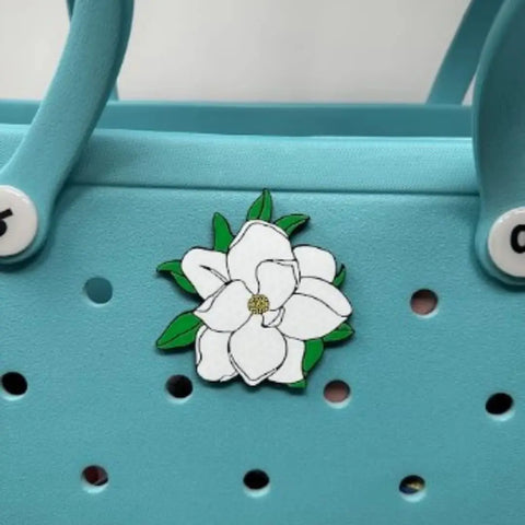 The Chatelet Manufacturing Baglets Magnolia Charm showcases a white magnolia flower with green leaves, perfect for decorating tote bags like Bogg Bags and Simply Southern bags.