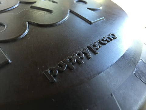 Close-up of a black SodaPup Magnum Bottle Top Flyer Ultra Durable Rubber Retrieving Frisbee (Large) with embossed text, crafted from magnum compound material; part of a number or letter appears in the background.