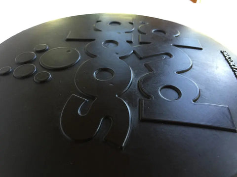 Close-up of the black Magnum Bottle Top Flyer Ultra Durable Rubber Retrieving Frisbee (Large) by SodaPup, showing its raised, embossed geometric shapes and numbers under natural light.