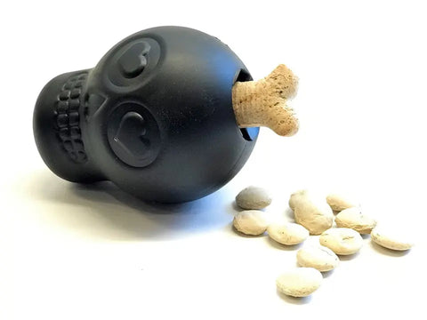 The SodaPup Magnum Sugar Skull EDispenser in black is an ultra-durable chew toy and treat dispenser shaped like a skull, shown on its side with a bone-shaped treat in its mouth beside light-colored dog treats on a white background.