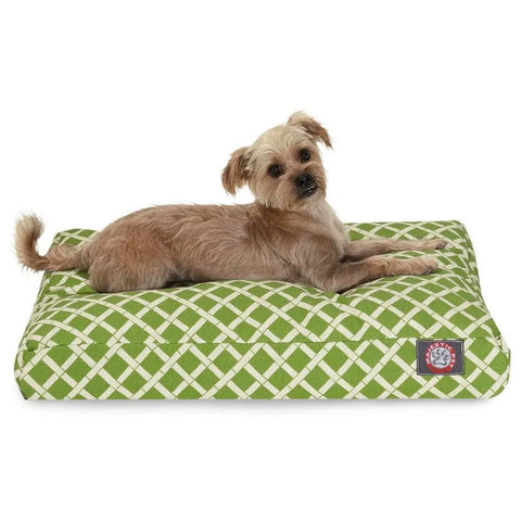 A small, light brown dog lies on the Majestic Pet Bamboo Memory Foam Dog Bed, featuring a green and white crisscross pattern and a red logo patch in the corner. The dog looks up with perked ears, enjoying the cozy, washable bed.