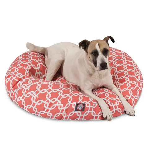 A white and brown dog relaxes on the Majestic Pet Links Round Dog Bed, which has a coral color, white geometric pattern, and a removable, washable cover. The pup lies with its front legs stretched out.