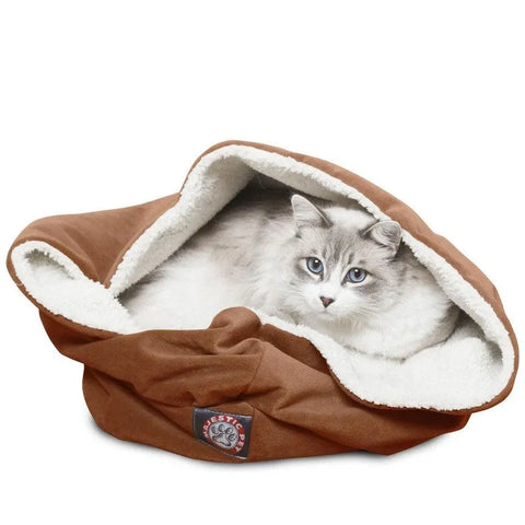 A fluffy, light gray cat with blue eyes is lying inside the Majestic Pet Suede Burrow Cat Bed. Its soft sherpa interior creates a cozy, partially enclosed space, while a paw logo patch decorates the front of this comfy bed.