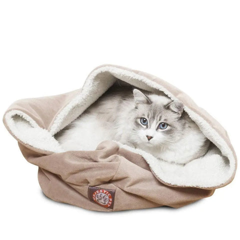 A fluffy white and gray cat with blue eyes relaxes in the Majestic Pet Suede Burrow Cat Bed, a soft tan pouch with a Sherpa interior, fleece lining, and a front patch featuring the brand’s paw print logo.