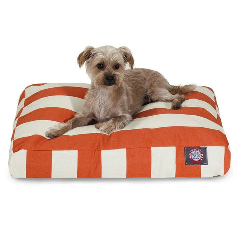 A small brown dog is lying on a Majestic Pet Vertical Stripe Memory Foam Dog Bed featuring wide orange and white stripes. The rectangular, washable cushion has a corner tag, and the dog appears alert yet relaxed.