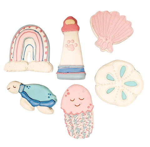 The Makapuu Box by Dogtown Bakery includes six pastel premium dog treats shaped like a rainbow, lighthouse, seashell, sea turtle, jellyfish, and sand dollar—decorated with pink, blue, and white icing on grain-free flour.