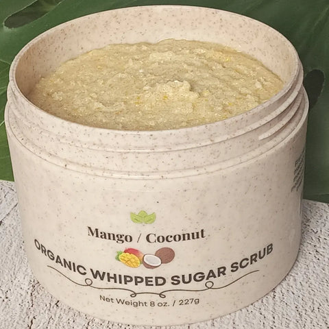 An 8 oz (227g) jar of Forev'Herbs Mango Coconut Organic Whipped Sugar Scrub, a vibrant yellow natural exfoliator, sits on a white surface with green leaves in the background.