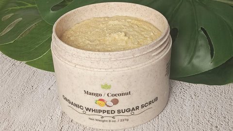An open jar of Forev'Herbs Mango Coconut Organic Whipped Sugar Scrub rests on a textured surface with green leaves behind it, showing the yellow scrub inside—an organic exfoliator for glowing, refreshed skin.