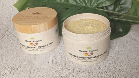 An open jar of Forev'Herbs Mango Coconut Organic Whipped Sugar Scrub sits on a textured surface beside its lid, with a green leaf in the background—a natural scrub for glowing, hydrated skin.