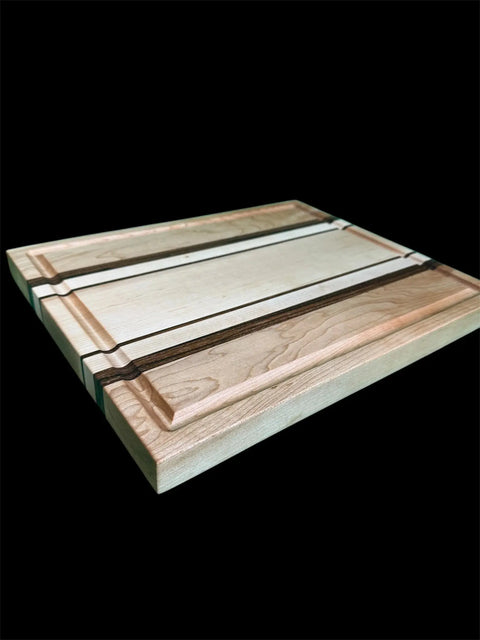 The Maple and Zebra Wood Inlay Cutting Board With Juice Groove by B. James Design & Woodwork, featuring light and dark stripes plus grooved edges, is shown against a black background.