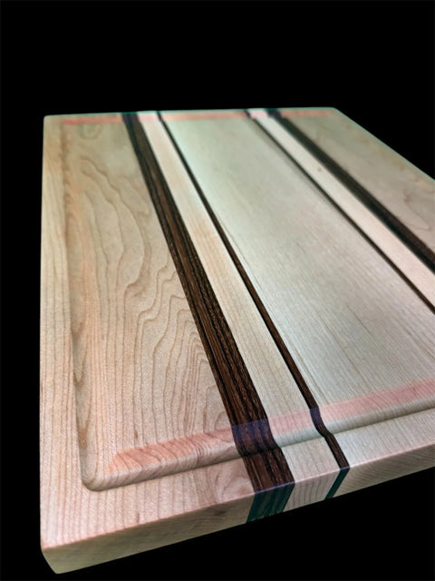 The Maple and Zebra Wood Inlay Cutting Board With Juice Groove by B. James Design & Woodwork, featuring striking light and dark grain patterns and parallel dark inlays, is displayed against a black background.