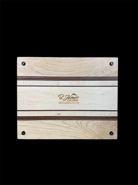The Maple and Zebra Wood Inlay Cutting Board With Juice Groove by B. James Design & Woodwork features dark and light wood stripes, four corner non-slip pads, and a centered engraved logo with website, set against a black background.
