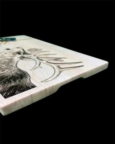 The Elk Epoxy Inlay Board by B. James Design & Woodwork is shown angled against a black background, featuring an engraved image of a stag’s head and antlers on solid maple.