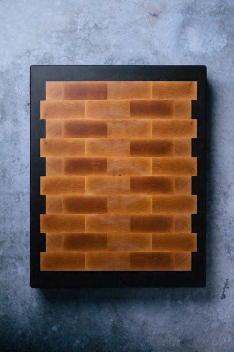 The Chef - End Grain Butcher Board - Maple by Montana Block features a rectangular maple design with a dark border and interlocking light brown blocks in a brick wall pattern, displayed on a textured gray surface.