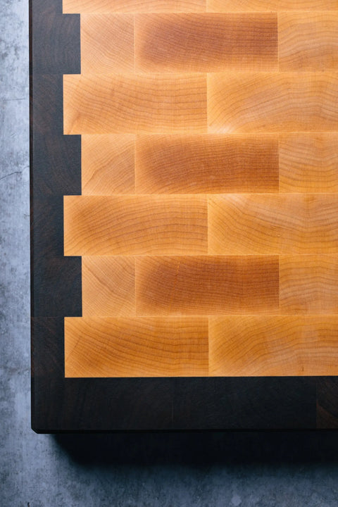 A close-up of The Chef – End Grain Butcher Board – Maple by Montana Block, an American-made butcher board with a checkerboard maple pattern and dovetail joints, featuring light and dark wood tones on a grey surface.