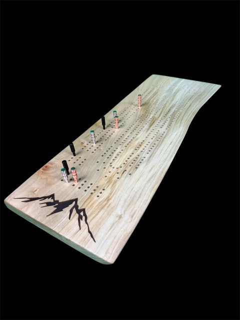 The Maple Live Edge Cribbage Board With Mountain Engraving by B. James Design & Woodwork features multiple rows of holes, colorful pegs, and a mountain engraving in one corner on a striking black background.