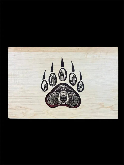 The Grizzly Inlay Serving Board by B. James Design & Woodwork features a large bear paw print with a bear’s face in the center pad and trees in the smaller pads, all handcrafted from solid maple against a black background.