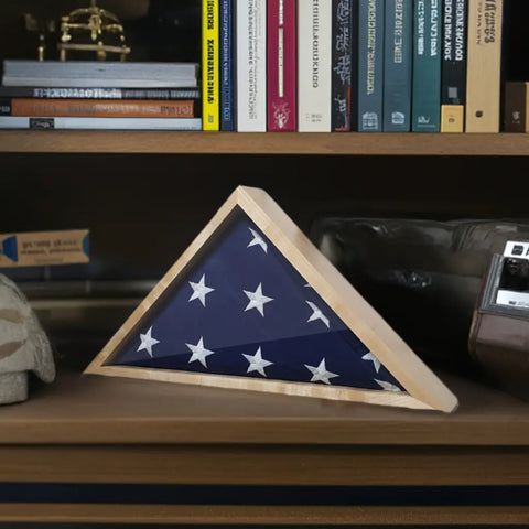 A folded American Flag rests in a Legacies of America Woodworking Company Maple 4x6 Flag Display Case on a wooden shelf, surrounded by books, a hat, and a vintage camera.