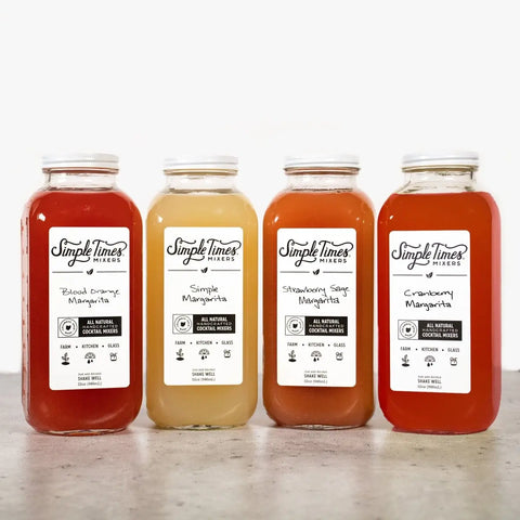 Four bottles from the Simple Times Mixers Margarita Bundle are displayed on a light surface. Flavors include Blood Orange Margarita, Simple Margarita, Strawberry Sage Margarita, and Cranberry Margarita.