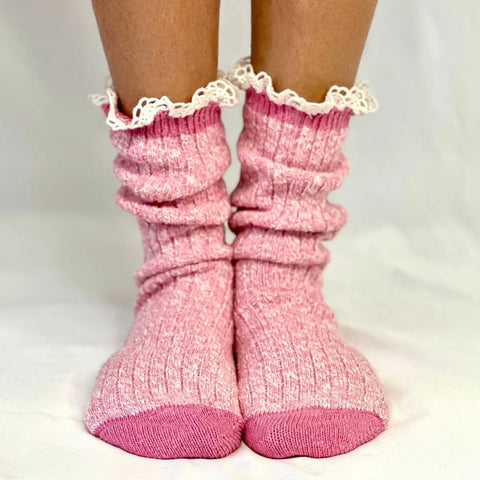 A pair of feet stand on a white surface wearing Catherine Cole MARLED Cotton Short Lace Boot Socks in pink, featuring thick ribbed fabric and ruffled lace trim at the top.