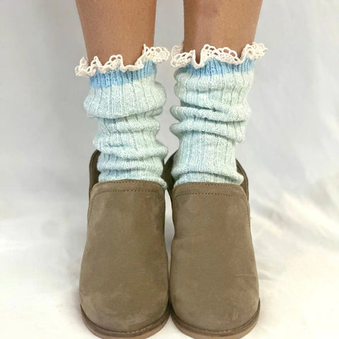 A pair of feet in Catherine Cole MARLED Cotton Short Lace Boot Socks in Carolina Blue, featuring delicate ruffled trim, styled with taupe suede slip-on shoes, standing on a white surface against a white background.