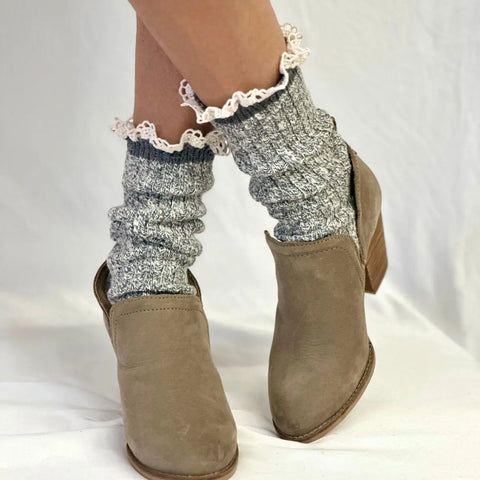 A close-up of a person wearing taupe ankle boots and Catherine Cole MARLED Cotton Short Lace Boot Socks in grey, with white lace trim scrunched above the boots, standing on a white surface.