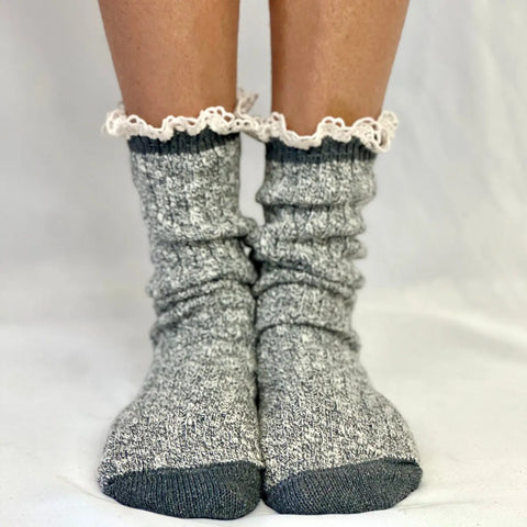 A pair of feet wear Catherine Cole MARLED Cotton Short Lace Boot Socks in grey, featuring dark toes and ruffled white lace trim at the top, standing on a light surface.