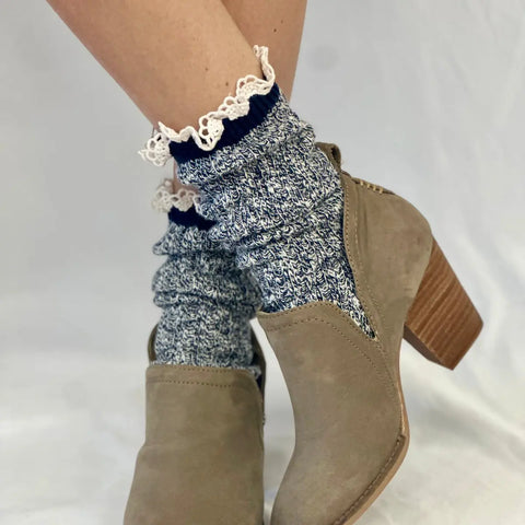 Close-up of a person’s legs wearing Catherine Cole MARLED Cotton Short Lace Boot Socks in navy with ruffled trim and taupe suede heeled ankle boots. The socks feature blue and white speckles, shown against a plain white background.