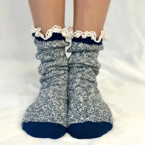 A close-up of feet in Catherine Coles MARLED Cotton Short Lace Boot Socks in navy, featuring blue and white marled cotton, navy toes, heels, and cuffs, with a frilly white lace trim at the top, standing on a white surface.