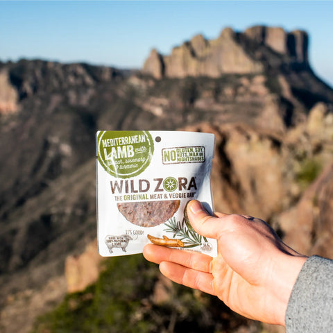 A hand holds a Wild Zora Bars - Mediterranean Lamb With Spinach, Rosemary & Turmeric 10-Pack, with mountains and rocky cliffs blurred in the background under a clear sky.