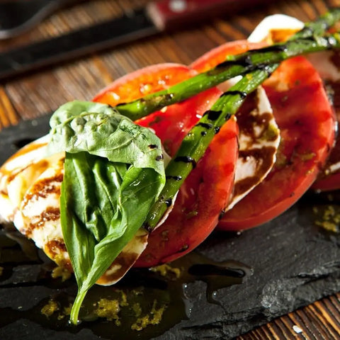 A close-up of Caprese salad with tomato, mozzarella, basil, grilled asparagus, balsamic glaze on slate, enhanced with Gustus Vitaes Taste of the Mediterranean - 3 Tins seasoning for an extra burst of flavor.