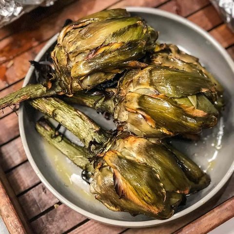 Three grilled artichokes with charred leaves sit on a gray plate atop wood, their browned edges enhanced by a sprinkle of Gustus Vitae’s Taste of the Mediterranean - 3 Tins Greek Tzatziki Sea Salt.