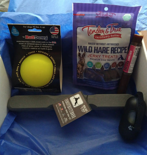 The Mega Max Indestructibone Gift Box by Bulletproof Pet Products Inc includes a durable chew, a waste bag holder, fetch stick, RuffDawg ball, and Tender & True Wild Hare Recipe jerky treats—all arranged on blue tissue paper. For dogs 51-100 lbs.