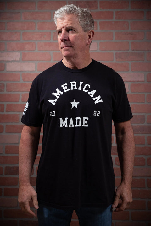 A gray-haired man stands before a brick wall wearing the Proudly USA American Made - Mens Cotton T-Shirt (Black), crafted from premium USA cotton. He looks slightly to the side with his arms relaxed at his sides.