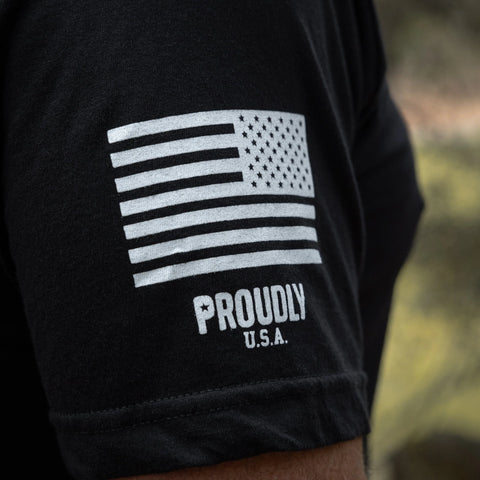 A close-up of the black American Made - Mens Cotton T-Shirt by Proudly USA shows a white stylized American flag above PROUDLY U.S.A. on the sleeve, with a blurred background.