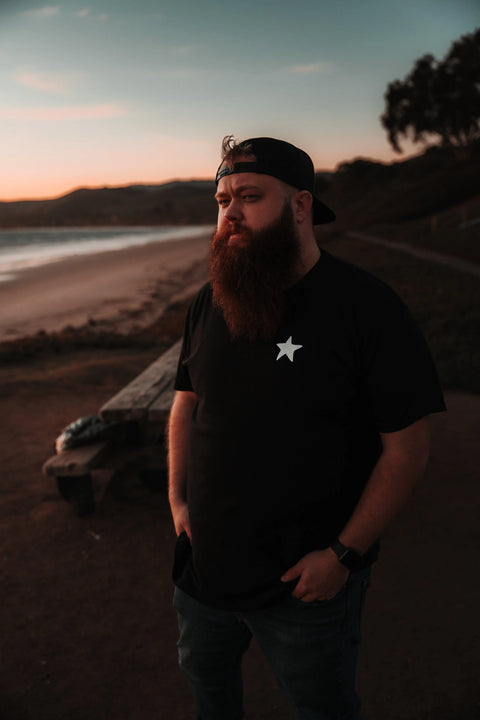 A bearded man in a Proudly USA Proudly Star Mens Cotton T-Shirt (Black), made from American grown cotton, with a backwards cap and watch, stands by the beach at sunset with hills and a picnic table in the background.