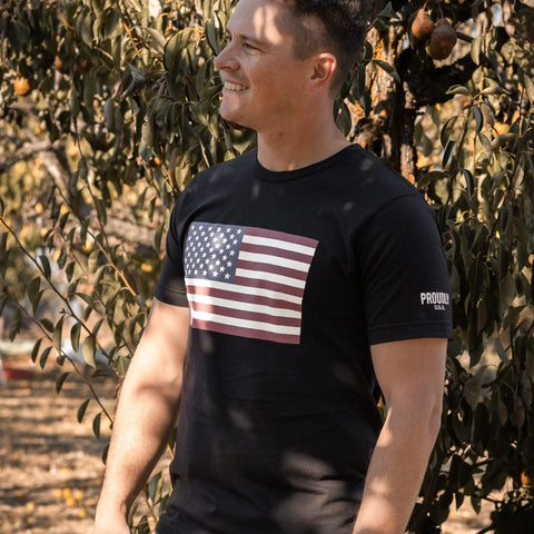 A smiling man stands outdoors near a tree in a Proudly USA American Flag - Mens Cotton T-Shirt (Black), sunlight filtering through the leaves in a warm, natural setting.