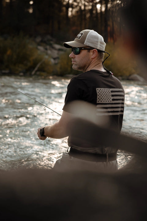 A man in sunglasses and a cap fishes by a river, wearing the Proudly USA Tattered Flag Mens Cotton T-Shirt (Black), surrounded by trees and rocks—embracing the outdoors in this classic patriotic shirt.